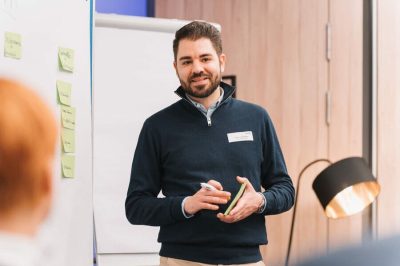 Video-Schulung Speaker Mann hält Schulung vor Flipchart, trägt dunklen Pullover, beige Hose; helles Zimmer, warme Lampenbeleuchtung.